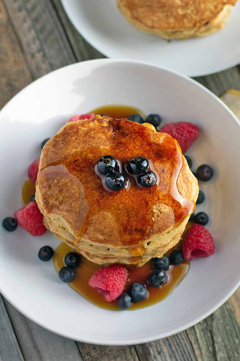 Vertical overhead image of a white plate of pancakes with fresh blueberries and raspberries, topped with maple syrup, with another plate of flapjacks at the top right of the frame, on an unfinished weathered wood surface.