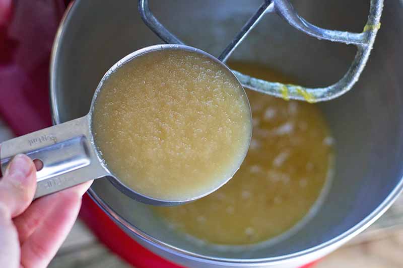 Horizontal image of a hand holding a metal measuring cup of apple sauce, about to pour it into the stainless steel bowl of a red stand mixer with paddle attachment.