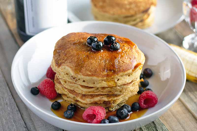 Horizontal image of a stack of pancakes in a shallow white ceramic bowl, with syrup and fresh berries, and another plate of flapjacks in the background, on an unfinished wood table.