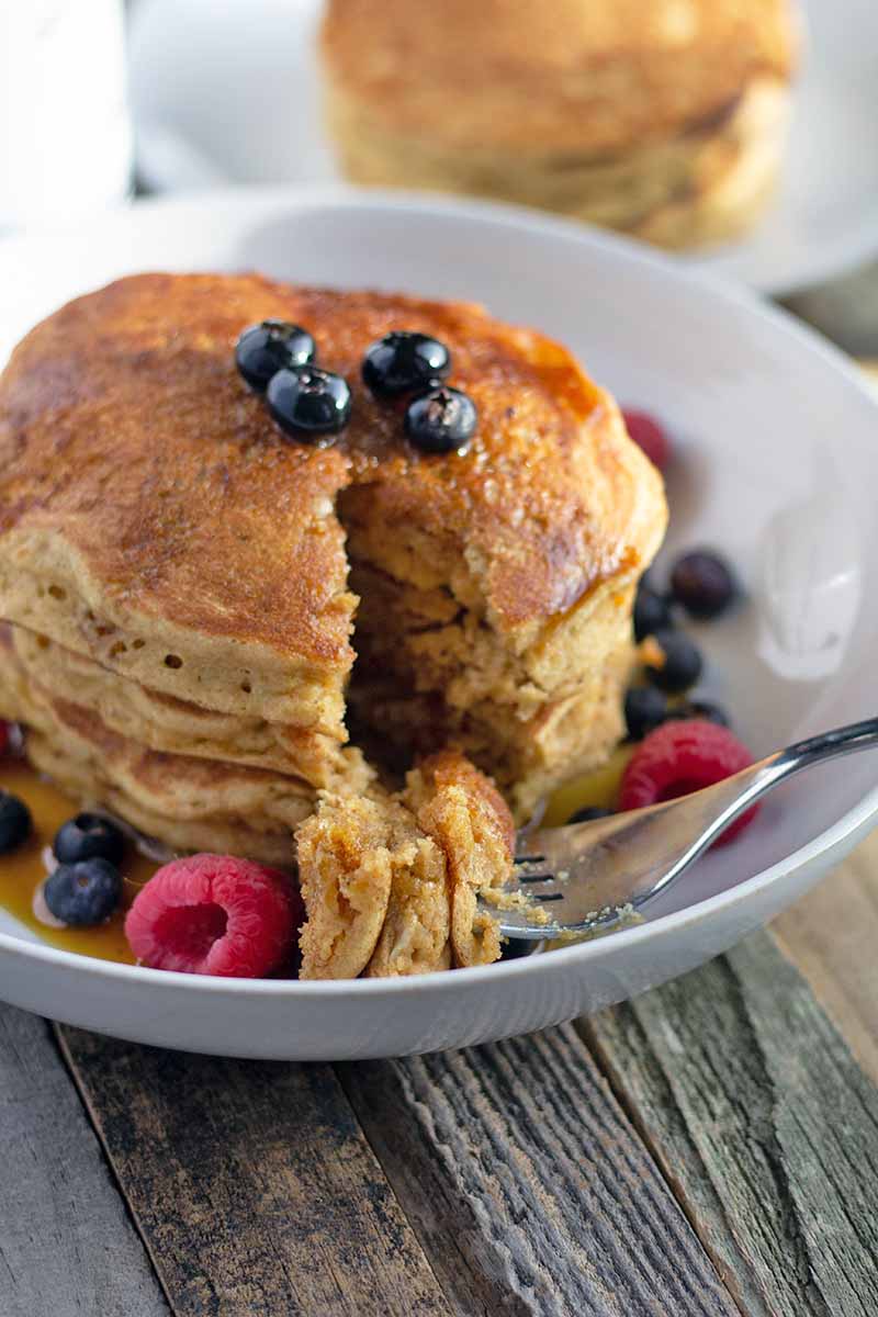 Vertical closely cropped oblique image of a stack of whole grain pancakes in a white shallow bowl with a piece cut out and sitting on a fork, with blueberries, raspberries, and maple syrup, and another stack of flapjacks on a plate in the background, on an unfinished wood surface.