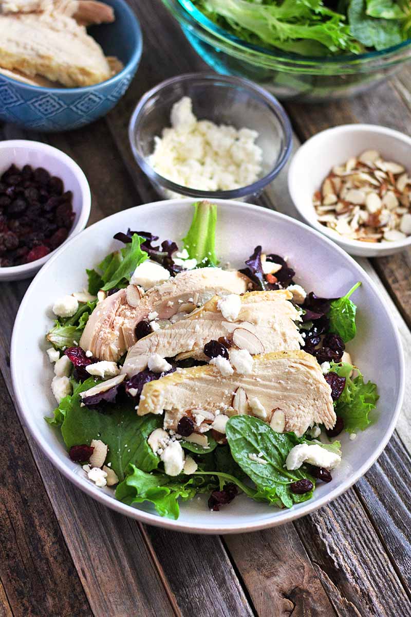 Vertical overhead image of a white bowl of sliced chicken breast on a bed of greens with crumbled goat cheese and dried fruit, surrounded by small bowls of more salad ingredients including slivered almonds, on an unfinished wood surface.