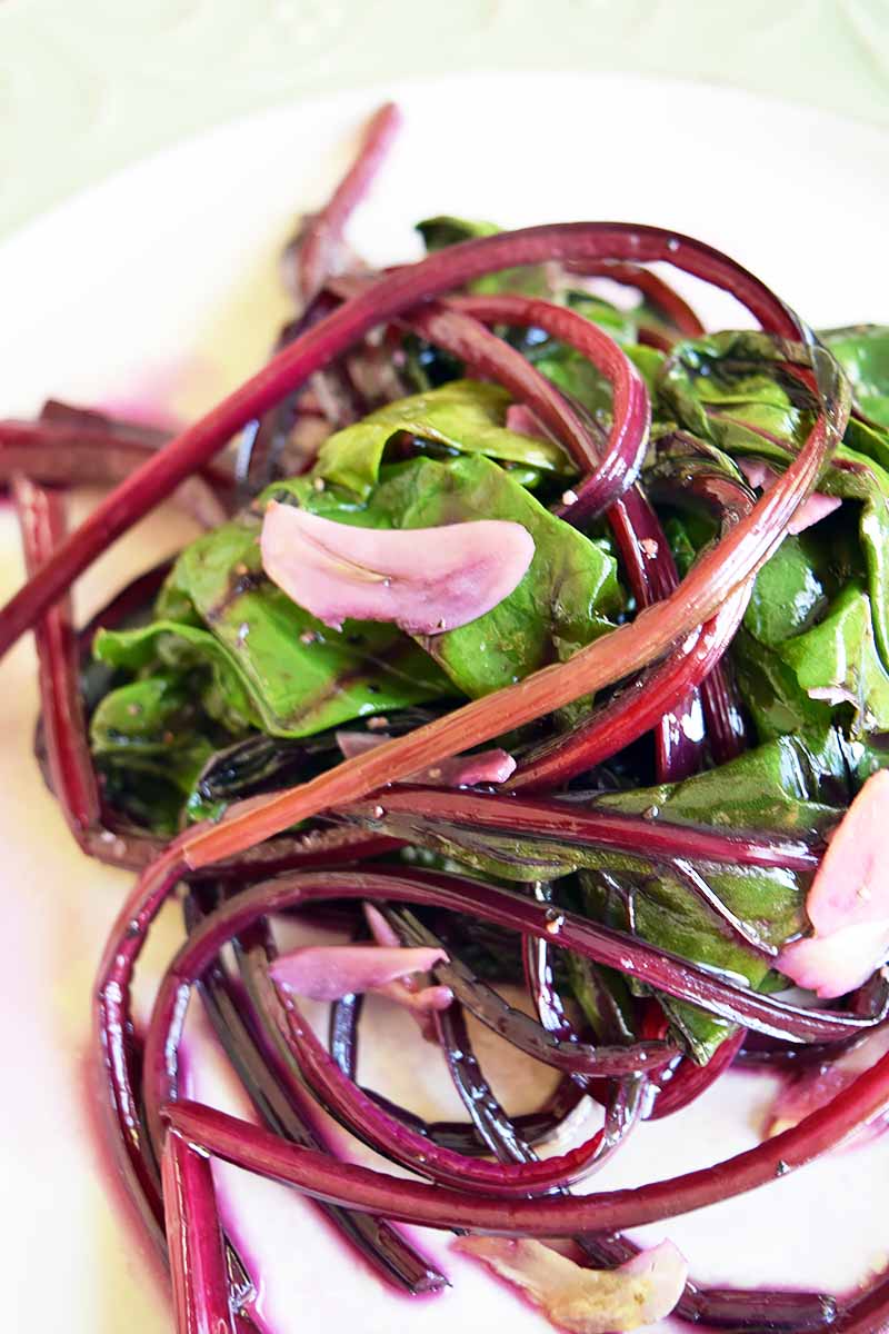 Vertical close-up image of a pile of wilted greens with purple stems and garlic slices on a white plate.