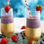 Horizontal image of a purple smoothie in two tall glasses with gold paper wrappers at the base of each, with red and white striped paper straws and a whole strawberry garnish, with scattered berries on a gray surface with a white cloth in the background, against a mottled blue and white backdrop.