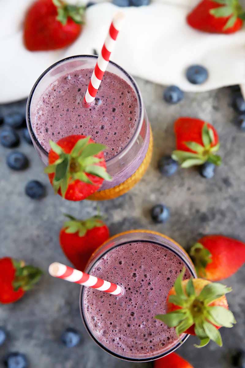 Vertical overhead image of a purple smoothie in two glasses, with berry garnish and red and white striped paper straws, on a gray surface with a white cloth at the top of the frame, and scattered fresh blueberries and strawberries.