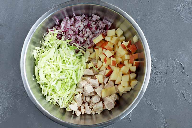 Horizontal overhead image of separate piles of shredded green cabbage, chopped purple onion, diced red apple, and chopped cooked chicken breast in a stainless steel bowl, on a gray surface.