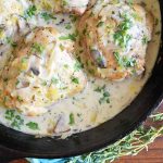 Overhead closely cropped horizontal image of chicken thighs in cream sauce with herbs in a black cast iron frying pan, on a brown wood surface with a sprig of thyme.