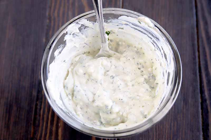 Horizontal top-down view of a small clear glass bowl of a white homemade goat cheese and cilantro crema with a spoon for stirring on a brown wood surface.