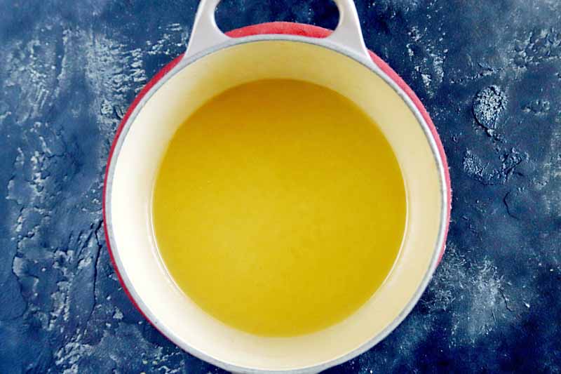 Overhead image of melted yellow butter in a cream-colored enameled saucepan with a red exterior, on a mottled blue background.