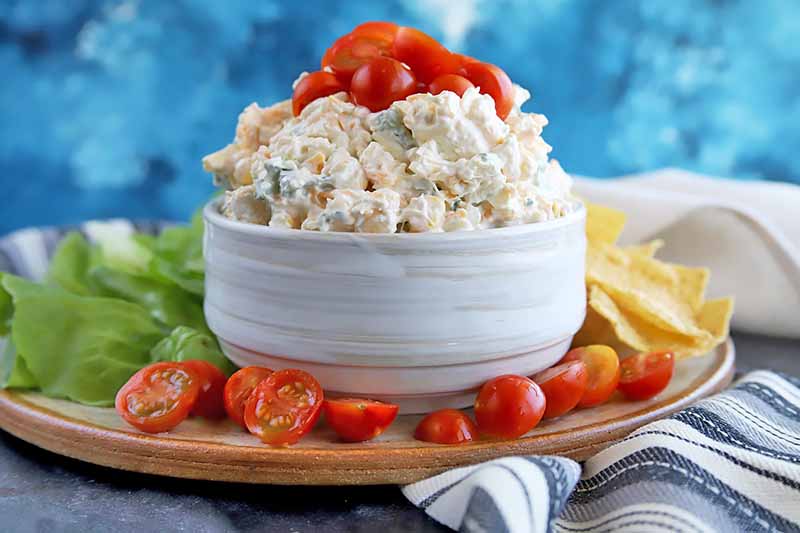 Horizontal image of a white bowl piled high with a creamy corn dip topped with tomatoes.
