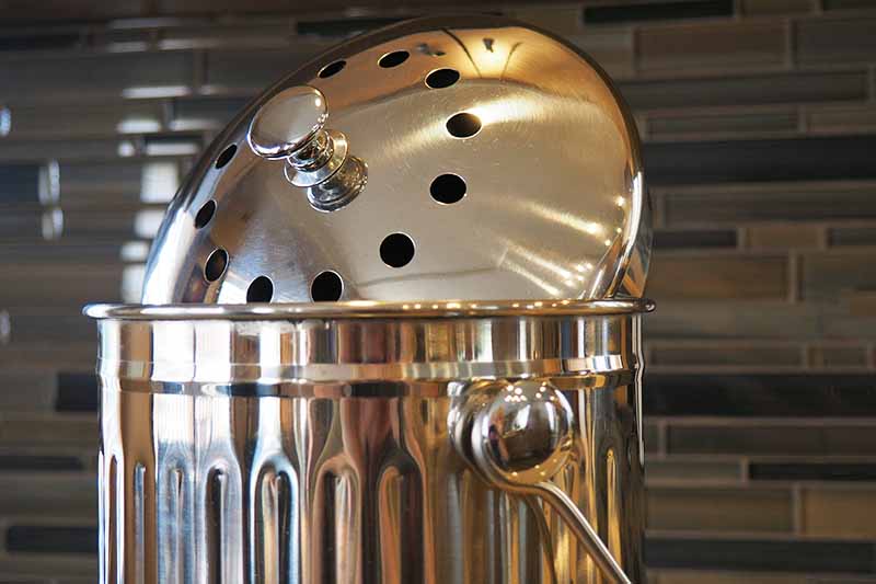 Horizontal head-on image of a stainless steel collection pail with a handle with a vented lid, with a brown tile wall in the background.