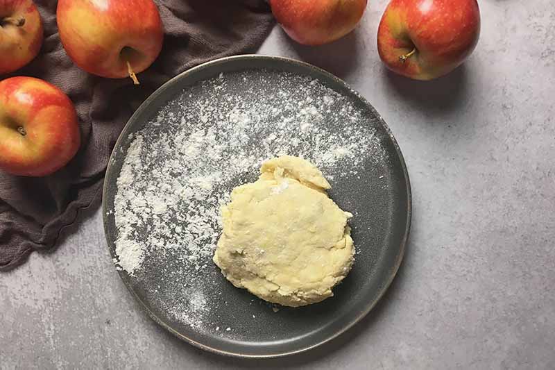 Horizontal image of a mound of dough on floured gray plate next to whole apples.