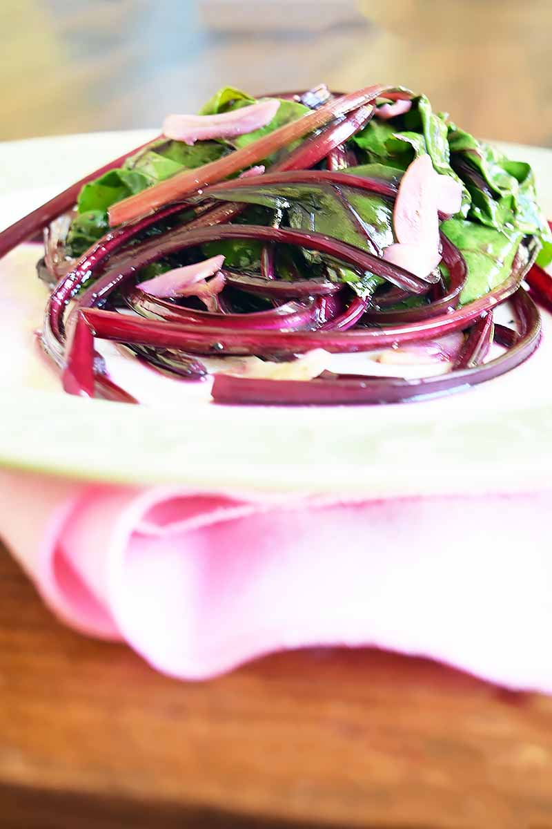 Vertical image of a pile of wilted green and purple vegetables on a white plate on top of a pink napkin.