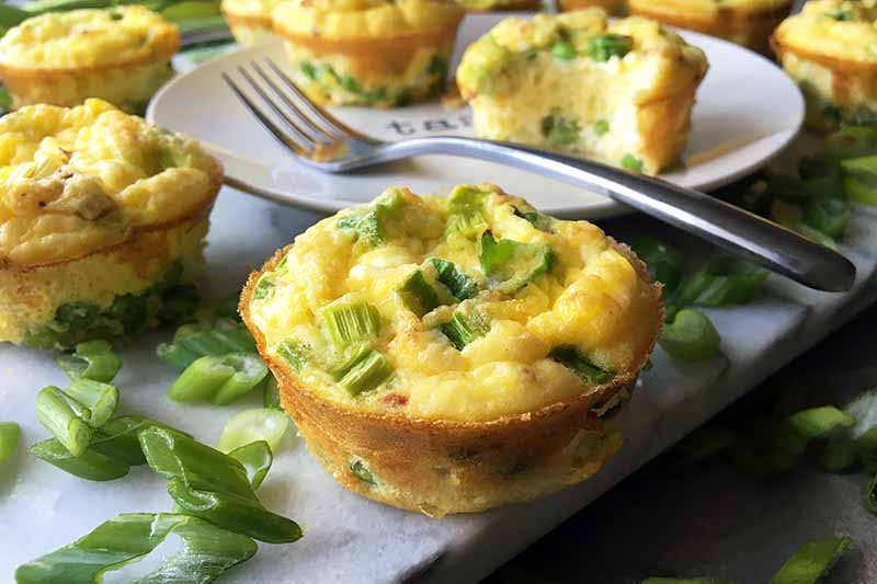 Horizontal image of mini frittata muffins, a white plate, a metal fork, and sliced green onions.