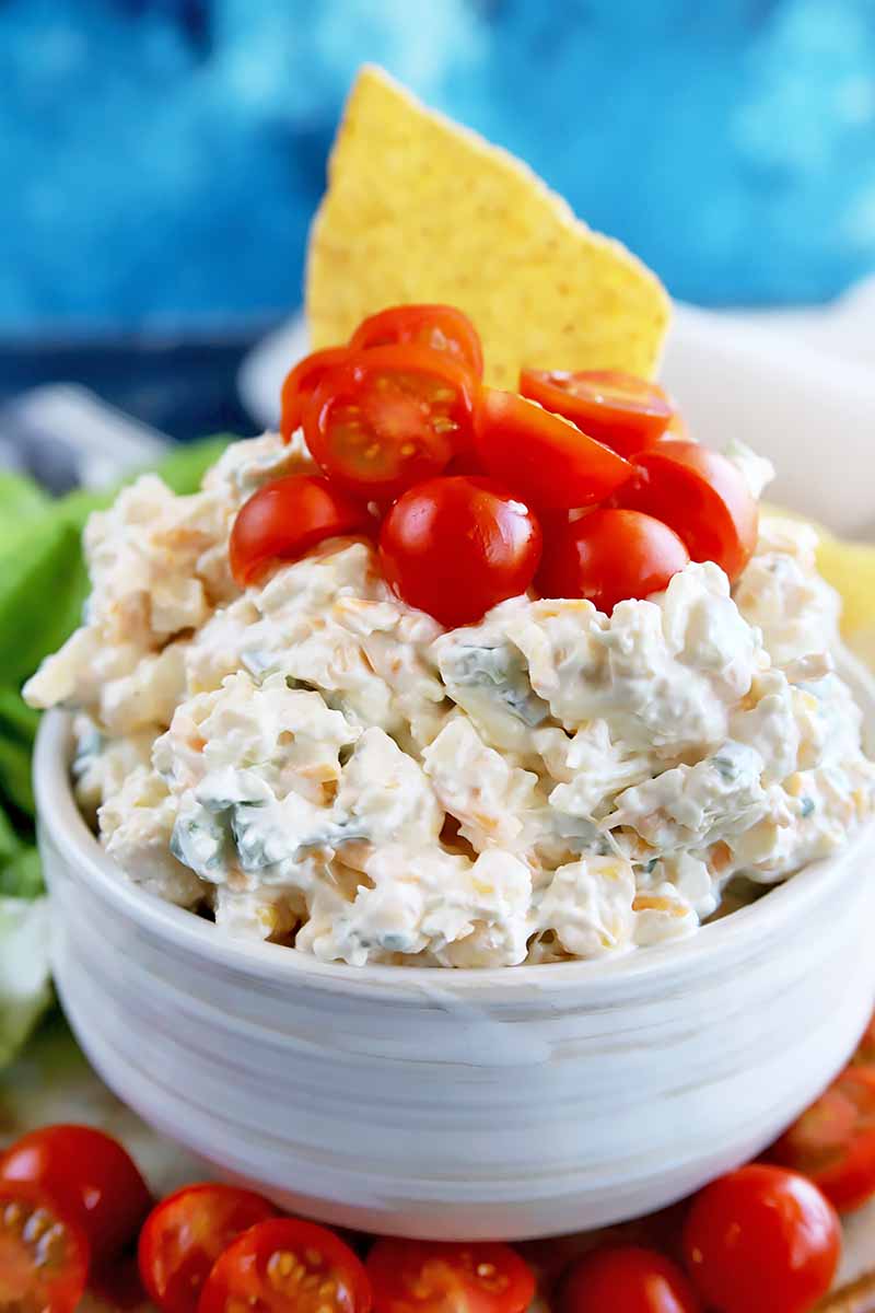 Vertical close-up image of a white bowl filled with a corn cream dip topped with sliced tomatoes and a tortilla chip with a blue background.