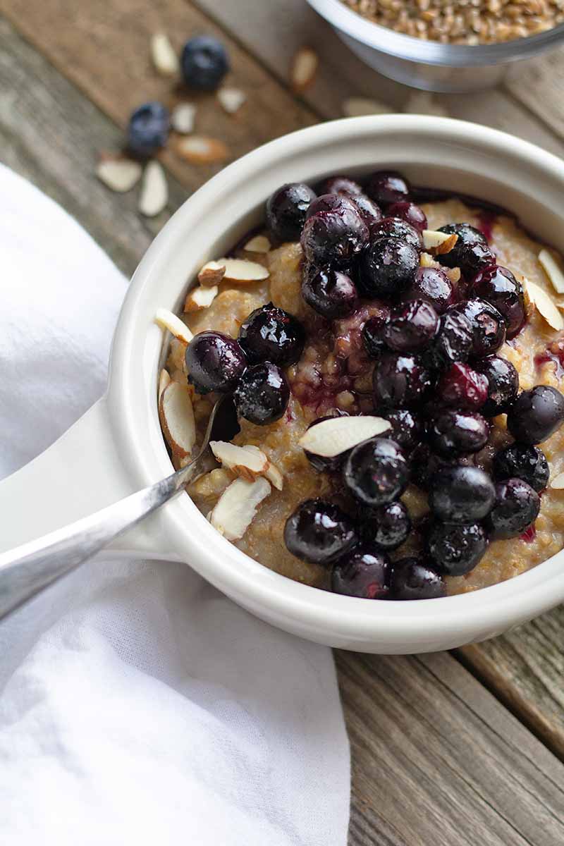 Overhead closely cropped vertical image of a white dish of hot breakfast cereal topped with cooked blueberries, with a white cloth and scattered fruit and slivered almonds, and a small glass dish of grain, on an unfinished wood surface.