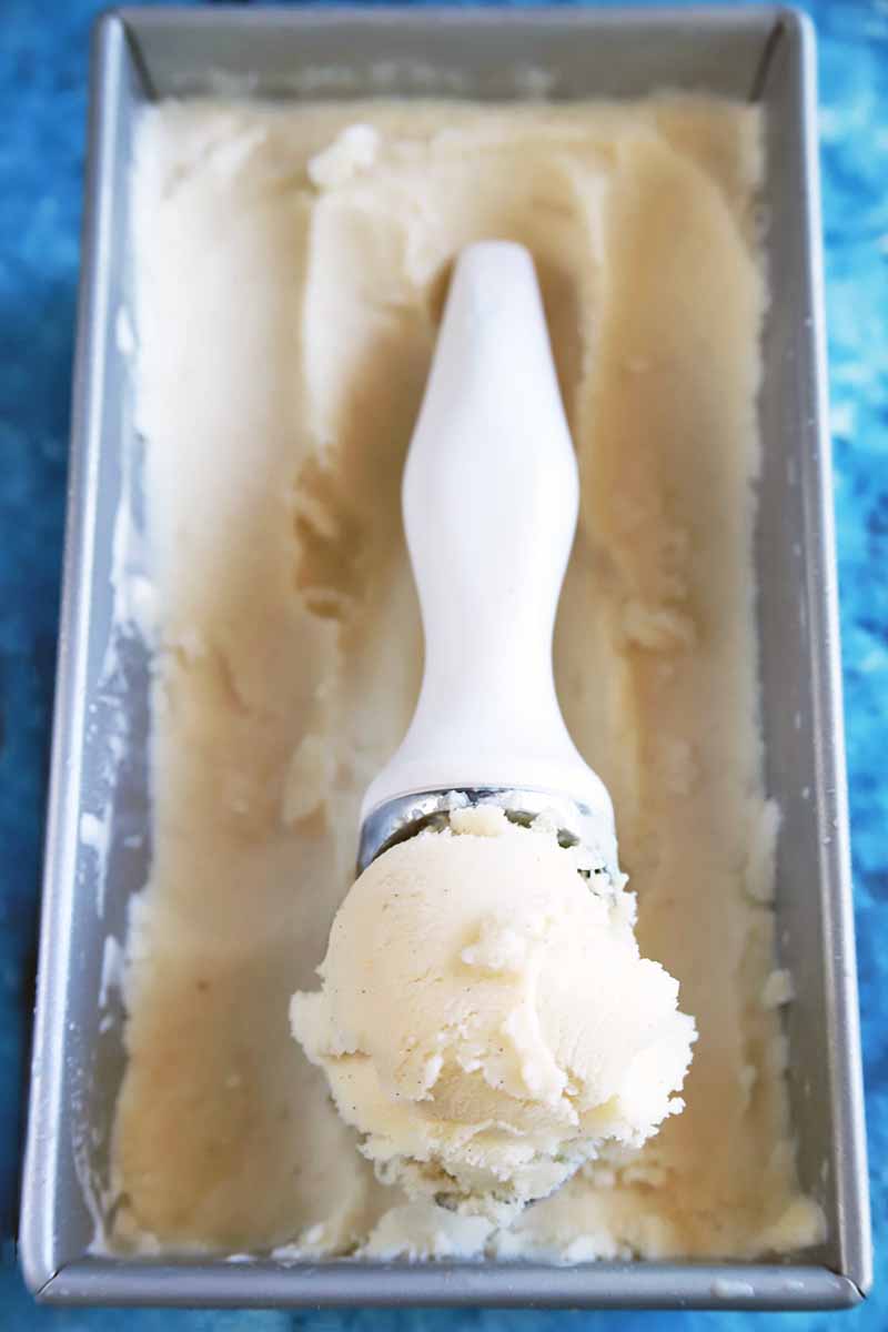 Vertical oblique overhead image of a metal loaf pan of vanilla ice cream with a white plastic and metal scooper on top being used to dish it out, on a blue and white speckled surface.