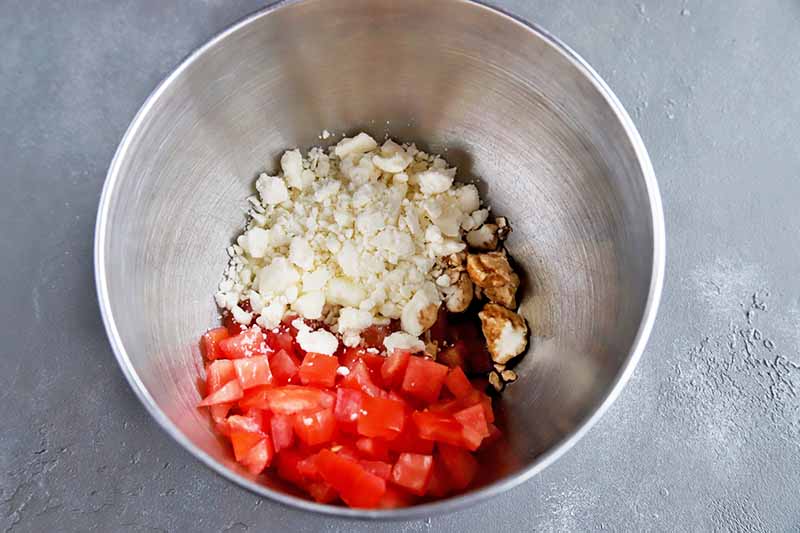 Horizontal image of chopped tomatoes, crumbled feta, and balsamic vinegar in a stainless steel bowl, on a gray surface.