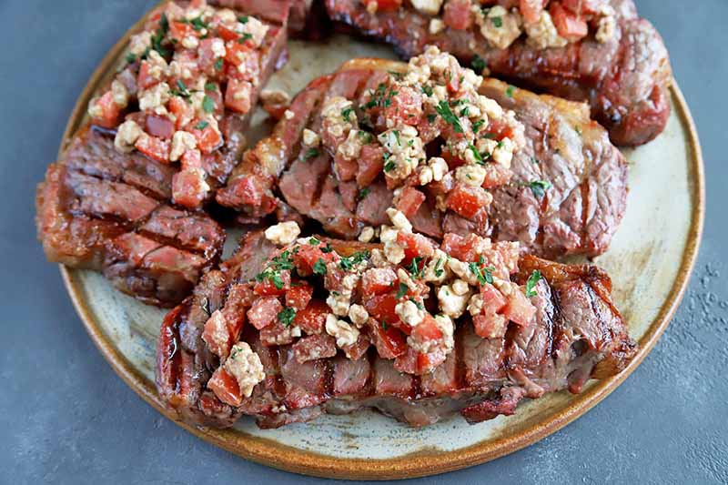 Horizontal oblique overhead image of a plate of grilled steak topped with chopped tomatoes and feta cheese, on a gray surface.