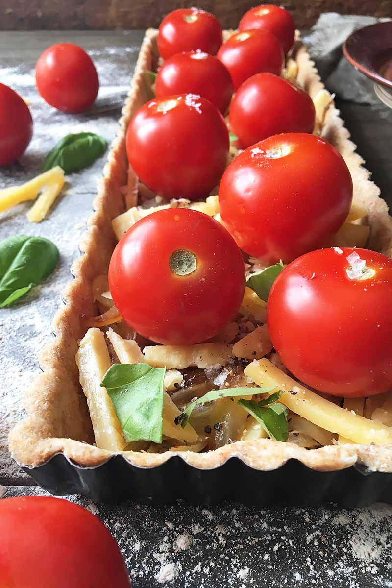 Vertical close-up image of an unbaked tart with whole tomatoes, torn basil, cheese, and onions.