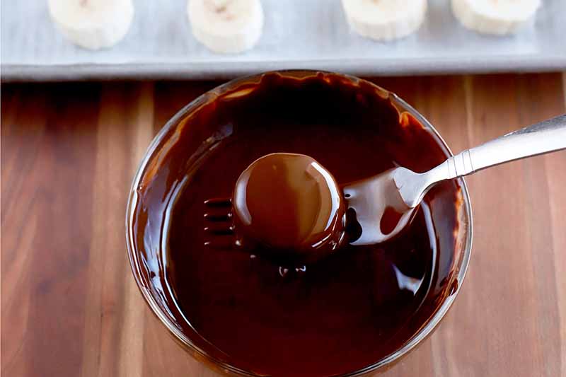 A fork holds a coated banana slice above a bowl of melted chocolate, allowing the excess to drip off, with a baking sheet of sliced fruit arranged in rows in the background, on a wood surface.