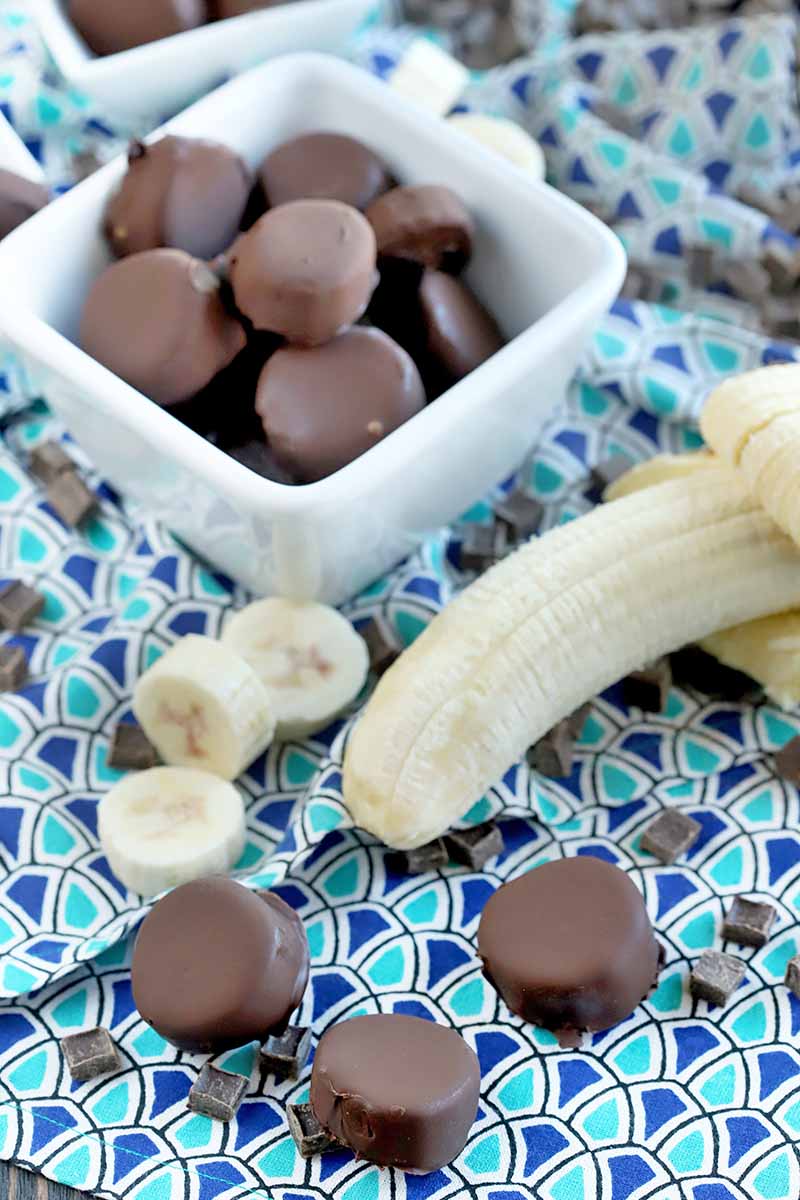 Vertical image of peeled and sliced banana with scattered chocolate chunks and frozen chocolate banana slices on a dark and light blue patterned cloth, with two white ceramic dishes of more of the dessert in the background.