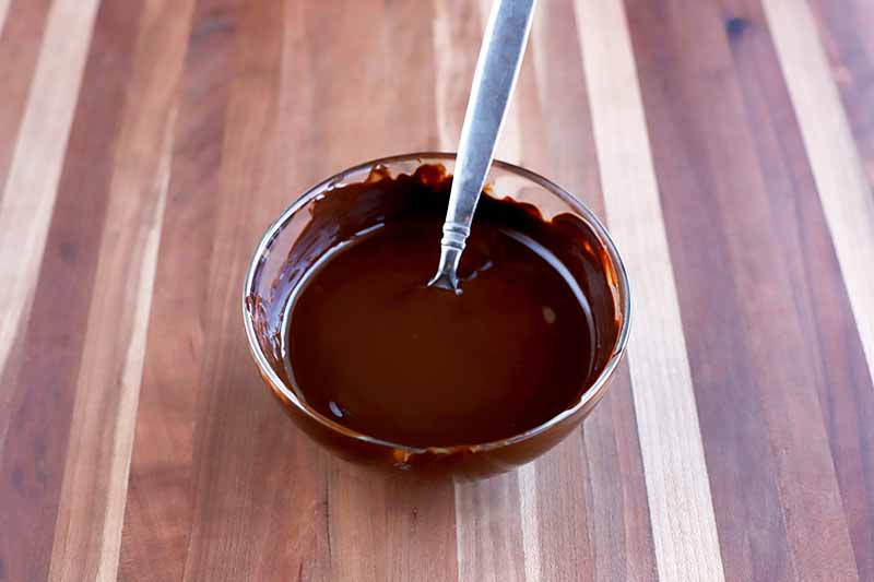 Horizontal image of a small glass bowl of melted chocolate with a spoon sticking out of it, on a beige and brown striped wood surface.