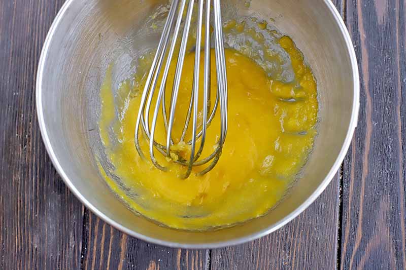 Horizontal image of a metal bowl with a whisk mixing a yellow mixture.
