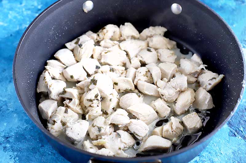 Marinated chopped pieces of boneless skinless chicken breast are cooking in a large nonstick frying pan, on a dark and light blue mottled background.