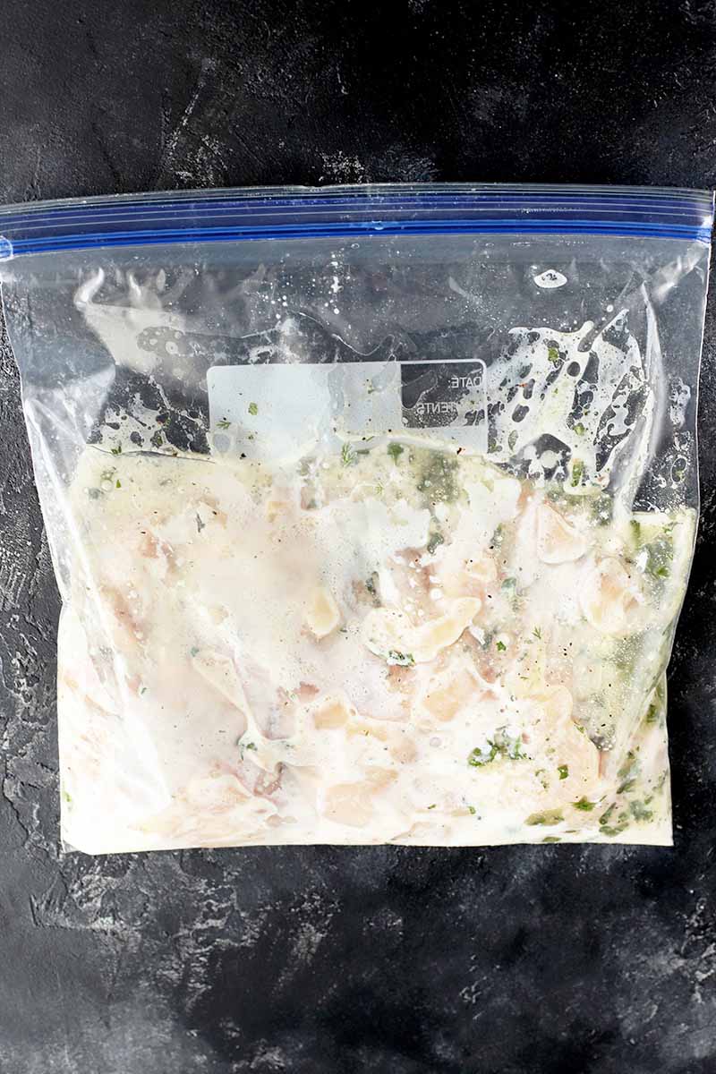 Overhead vertical image of a clear plastic zip-top bag with a blue plastic zipper, filled with a white marinade and pieces of raw chicken breast, resting flat on a dark gray surface.