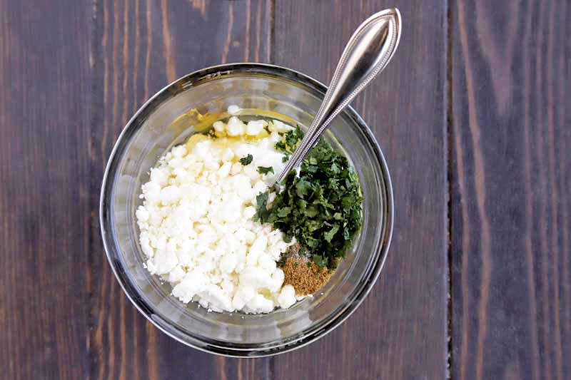Horizontal overhead image of a small clear glass bowl of crumbled goat cheese, chopped cilantro, and spices, with a spoon for stirring the mixture, on a brown wood surface.