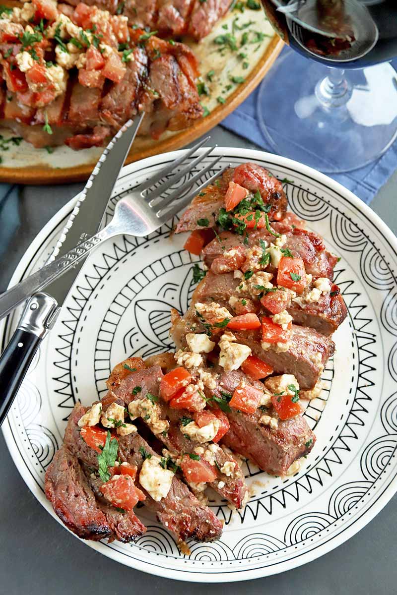 Vertical overhead image of a white plate with a black pattern, with a sliced portion of grilled New York strip steak topped with crumbled feta, chopped tomatoes, and herbs, with a knife and fork on the rim of the dish, and a serving platter of more beef in the background, with a glass of red wine at the top right of the frame, on a gray surface with a blue cloth.