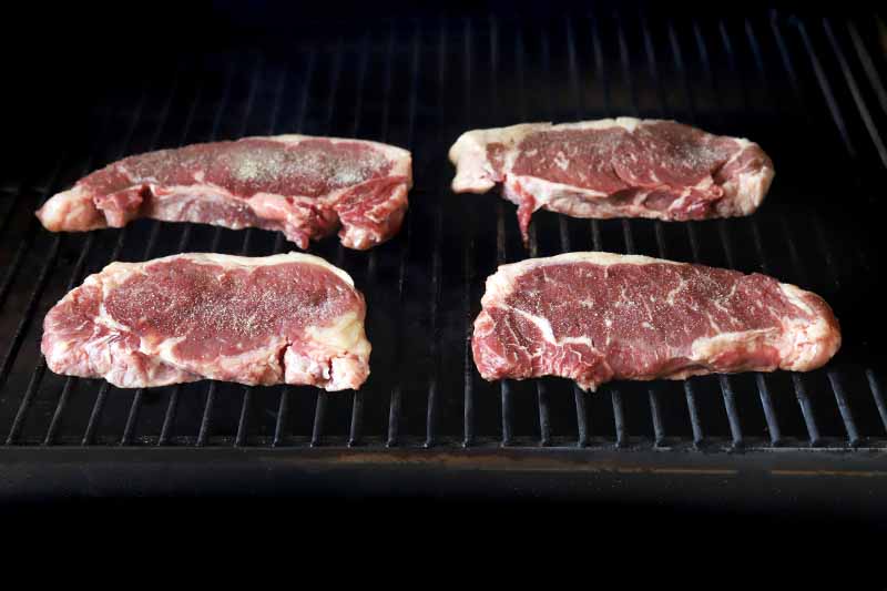 Four steaks grilling on metal grates, with a black background.