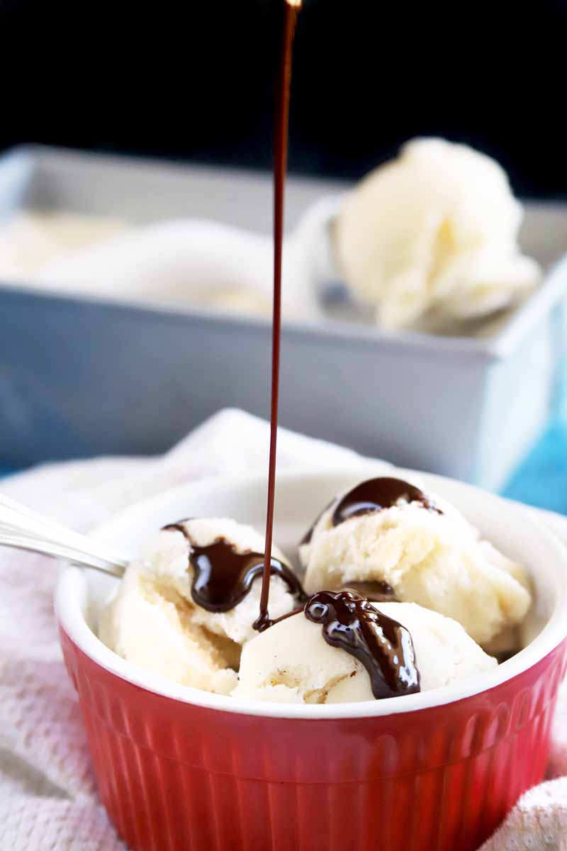 Vertical image of chocolate syrup being drizzled on top of three scoops of vanilla ice cream in a red and white ceramic ramekin, with more of the frozen dessert in a metal loaf pan in the background topped with a white plastic and metal scooper being used to dish it out, on a white kitchen towel with a black background on a blue surface.