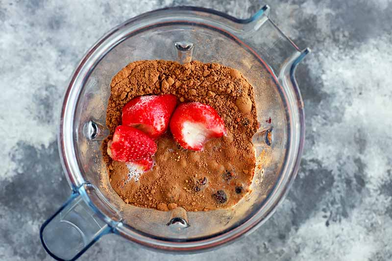 Horizontal overhead image of a clear plastic blender pitcher filled with a liquid topped with cocoa powder and whole hulled strawberries.
