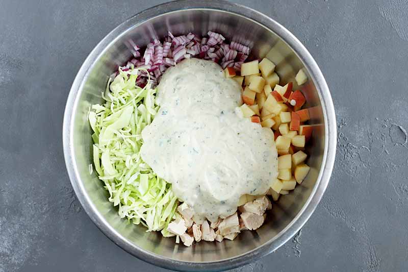 Horizontal overhead image of a stainless steel bowl filled with separate piles of shredded green cabbage, chopped purple onion, diced apple with the peel on, and chopped cooked chicken breast, with a blob of yogurt dressing in the center.