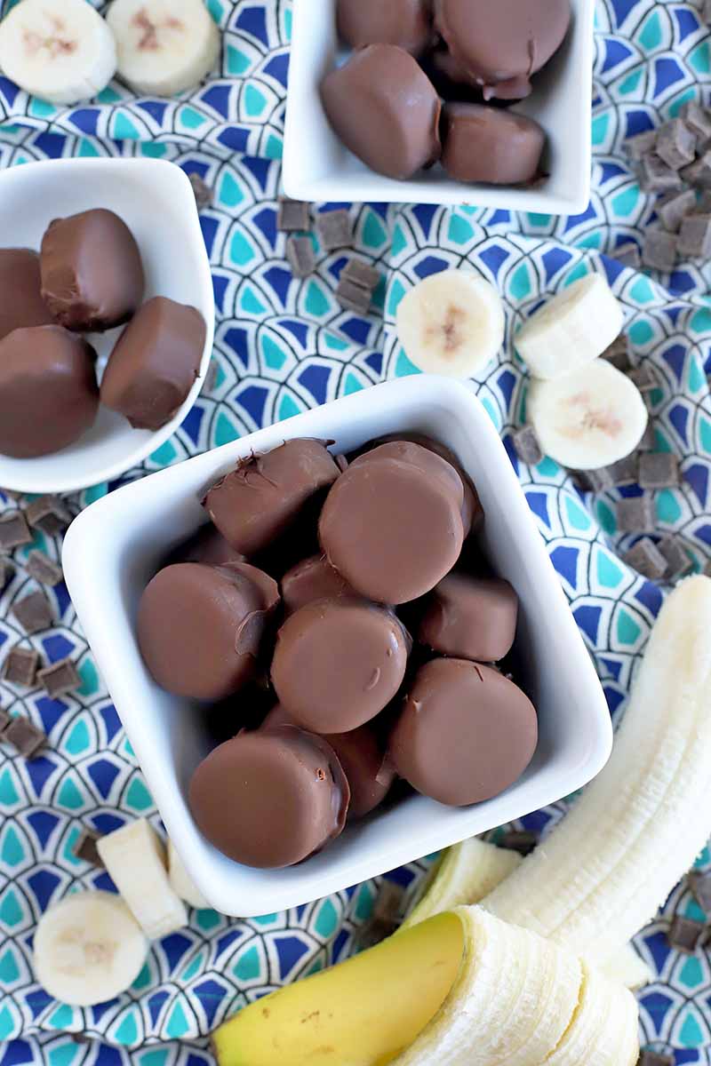 Vertical overhead image of one medium and two smaller white ceramic bowls of chocolate covered banana bites, on a dark and light blue patterned cloth with scattered banana slices, candy bits, and a half-peeled ripe banana.