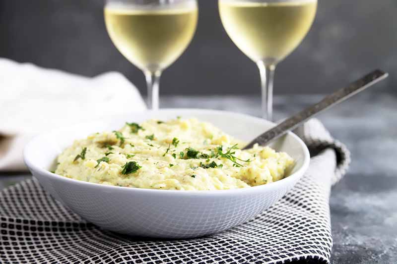Horizontal image of a white bowl filled with creamy mashed spuds on a checkered towel in front of two glasses with white wine.