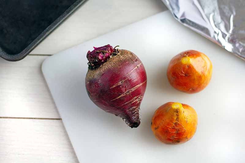 Horizontal overhead image of one medium-sized red beet and two smaller golden beets on a white plastic cutting board, on a whtie painted wood surface with aluminum foil and a metal baking pan at the top right and left corners of the frame.