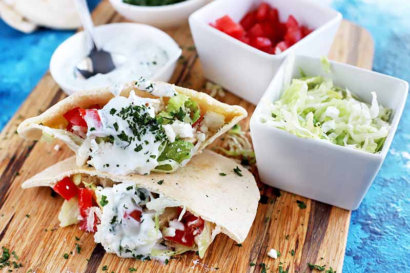 Hoizontal image of two Greek-style chicken pitas with fresh vegetables, herbs, and a white sauce, on a wooden cutting board with round and square dishes of various sizes filling with extra topping ingredients, on a blue cloth surface.