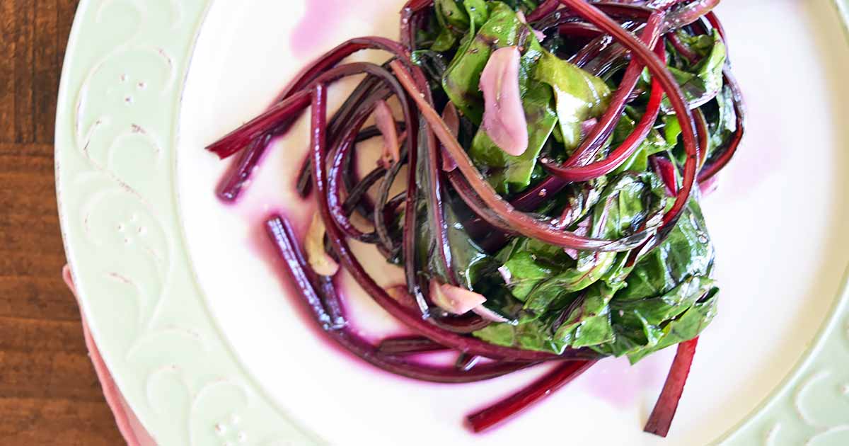 Horizontal image of wilted vegetables with purple stems on a white plate on a wooden table.