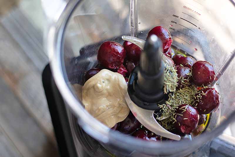 Closely cropped horizontal oblique overhead image of pitted fresh cherries, seasonings, and a dollop of Dijon mustard in a food processor, on a wood surface.