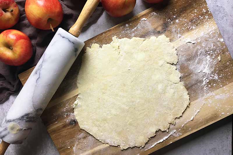 Horizontal image of a rolled out section of dough next to a rolling pin on a wooden cutting board.