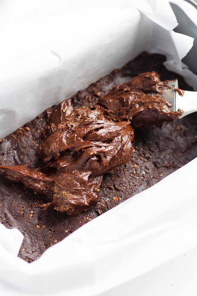 Vertical image of dollops of thick chocolate frosting on top of a dessert in a pan lined with parchment paper.