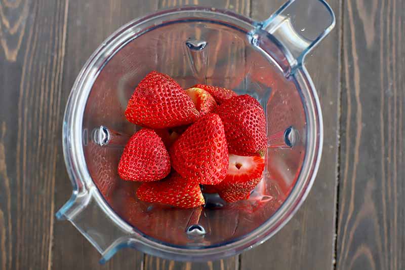 Horizontal image of a blender with a pile of fresh strawberries.