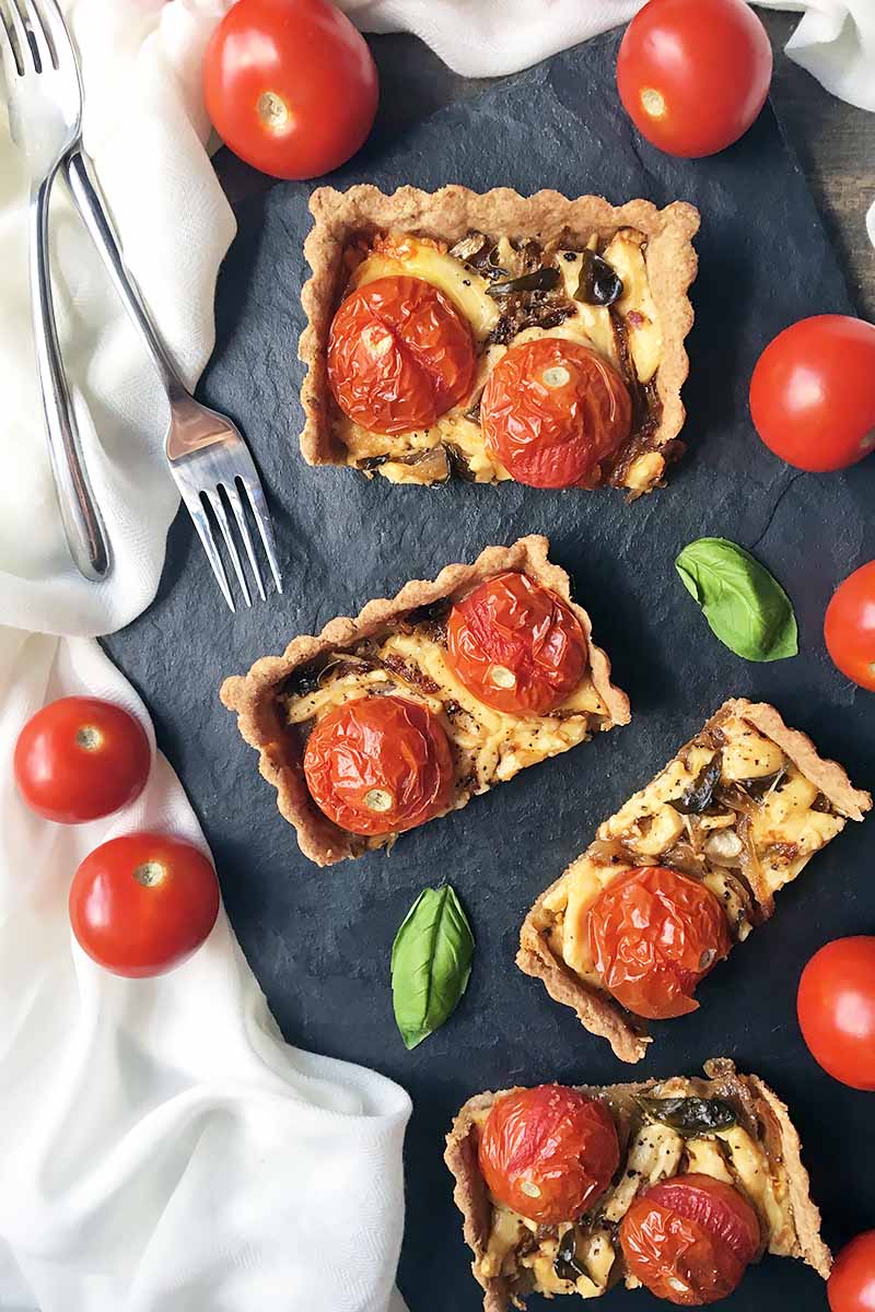 Vertical image of 4 cut pieces of a rectangular pastry topped with blistered tomatoes next to a white towel, tomatoes, and forks.