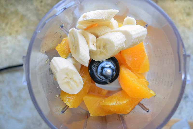 Horizontal overhead image of sliced banana and orange supremes in a blender with a clear plastic canister, on a kitchen countertop.