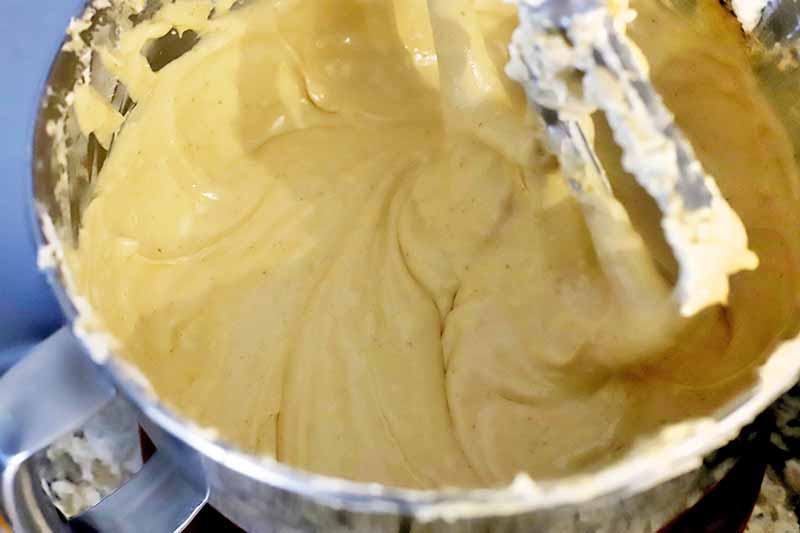 Horizontal closely cropped image of a creamy yellow mixture in a stainless steel mixing bowl, with a paddle attachment stirring it.