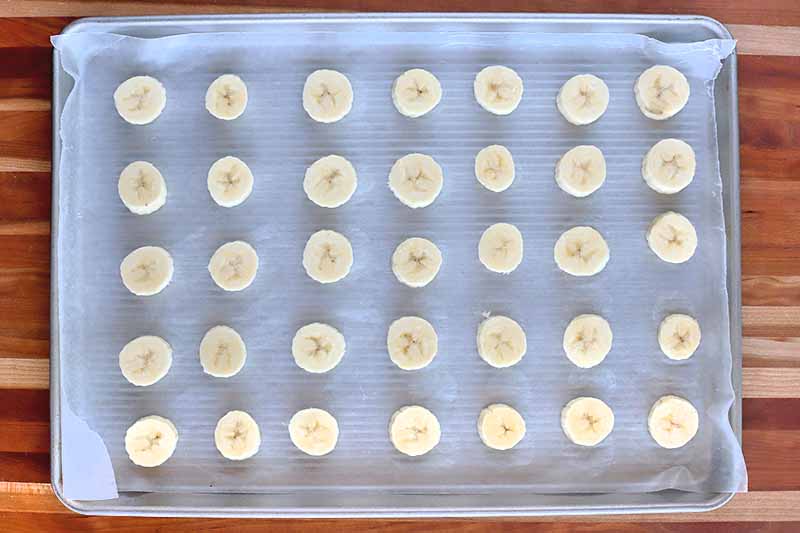 Horizontal overhead image of a baking sheet with 35 banana slices arranged in five rows, on a brown striped wood surface.