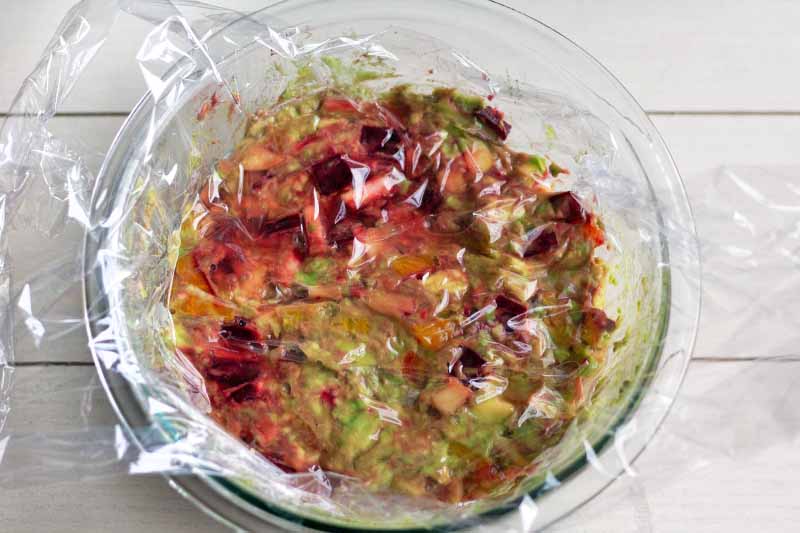 Overhead image of beet, apple, and avocado guacamole in a glass bowl, topped with plastic wrap and some water, on a white painted wood surface.