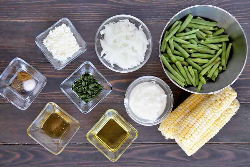 Overhead horizontal image of small round and square glass bowls of crumbled goat cheese, sliced onions, yogurt, oil, chopped herbs, spiced, and salt and pepper, with a larger stainless steel bowl of green beans and three husked ears of corn to the right, on a brown wood surface.